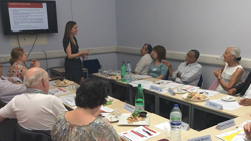 A group of laypersons sit in a U-shape around tables, conversing with a researcher who presents from a screen at the front of the image. The researcher is female with a short-sleeved black and white spotted dress,